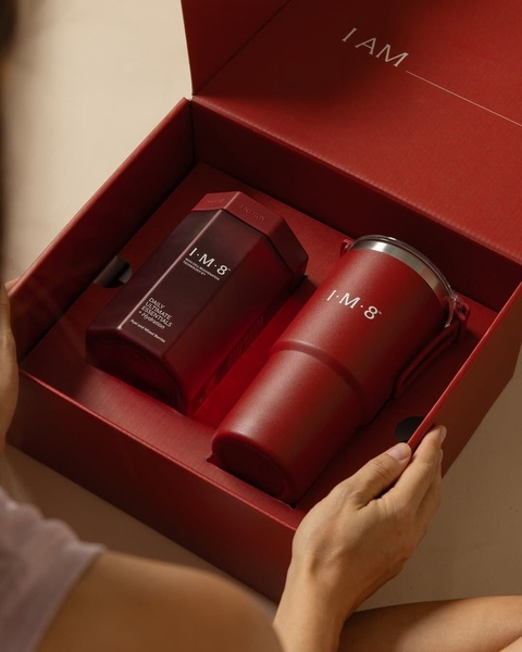 A high-angle shot of a person opening a premium red gift box containing an energy supplement jar and a matching insulated tumbler.